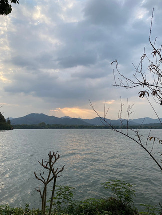 Calm lake with rippling water, surrounded by silhouetted trees. Mountains under a cloudy sky with a hint of sunset glow. Peaceful mood.