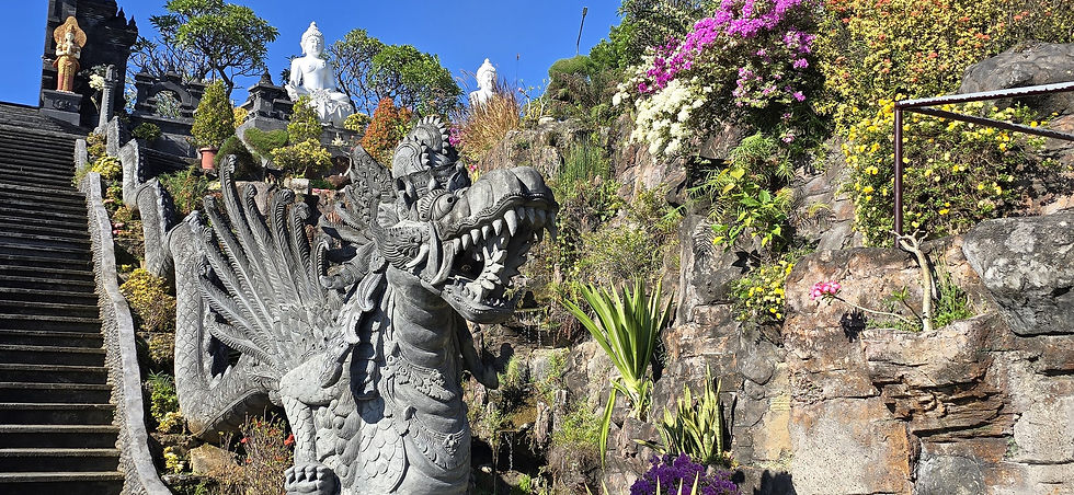 Stone dragon statue beside temple steps, with Buddha figures, bright flowers and blue sky in a lush hillside garden.