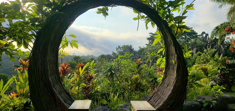 Circular wooden structure with two benches set in a lush tropical garden. Vibrant green foliage and colorful plants surround, under a cloudy sky.