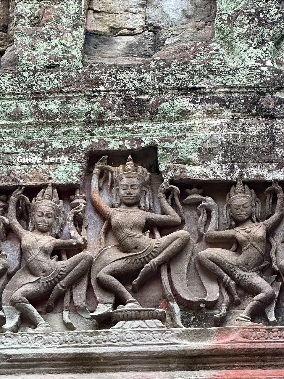 Stone carving of three dancing figures on an ancient temple wall, adorned with detailed patterns. Weathered texture and moss visible.