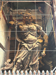 Statue of a fierce guardian deity in a temple, behind netting. The figure is muscular, wearing flowing robes, and holding a weapon.