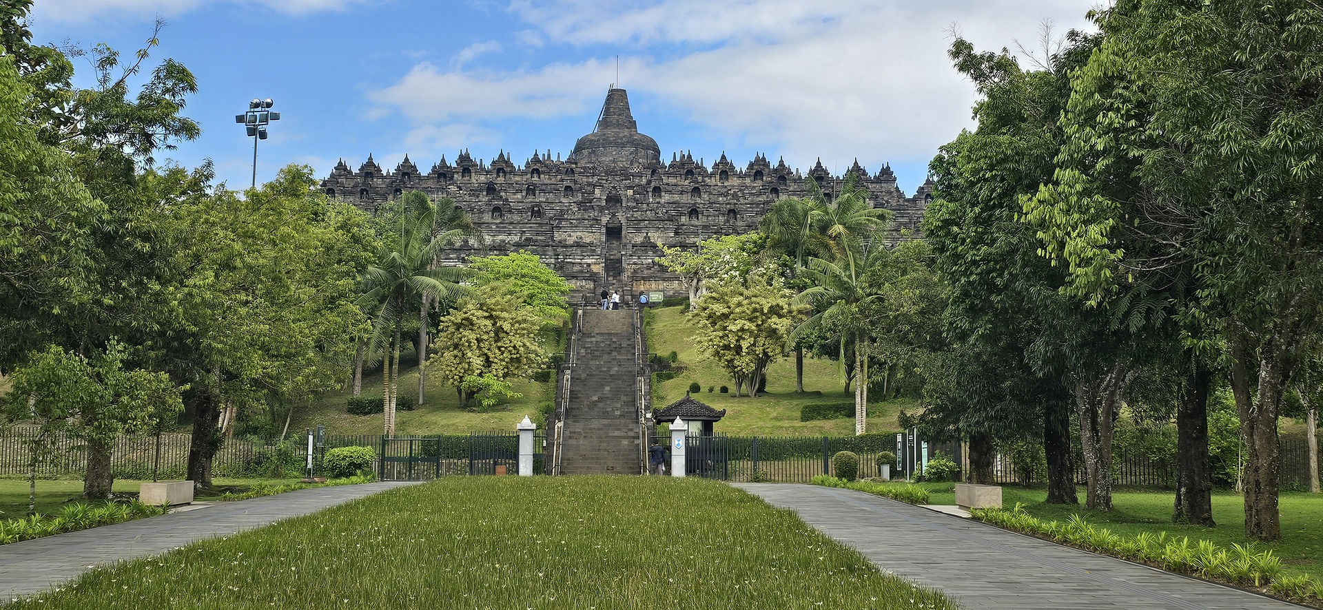 Ancient stone temple with intricate carvings under a blue sky, surrounded by lush green trees. The scene is serene and majestic.