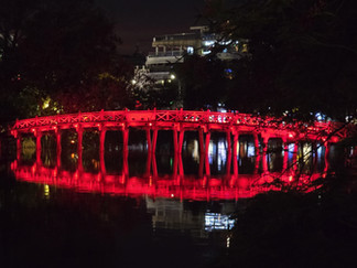 A red-lit wooden bridge reflects on a lake at night, with trees and buildings illuminated in the background, creating a serene scene.