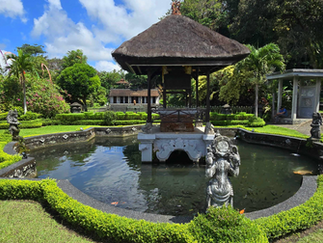 Lush garden scene at Pura Gangga Temple with a central pond, ornate statue, and traditional pavilion. Vibrant greenery surrounds under a blue sky with clouds.