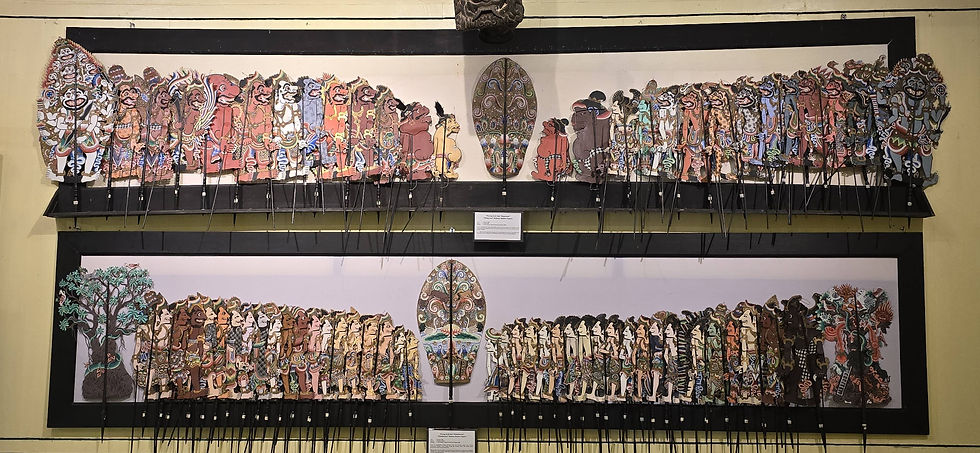 Colourful shadow puppets displayed on a two-tiered wooden frame with intricate designs, set against a beige wall.