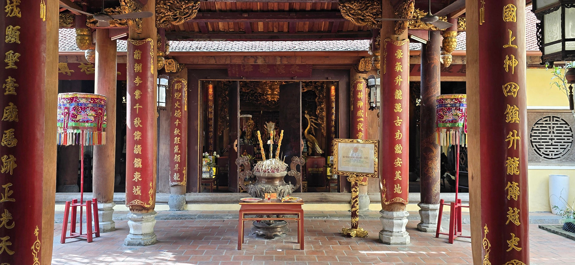 Ornate temple interior with red columns featuring gold text. A decorated parasol and incense burner are in front, creating a serene ambiance.