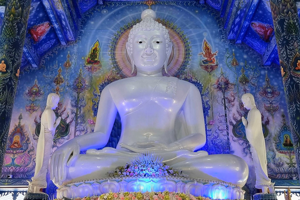 Large white Buddha statue in a colorful temple interior, flanked by two standing figures. Vibrant blue hues and intricate patterns adorn the walls.