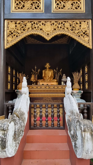 A golden Buddha statue sits in an ornate gold and black alcove, with carved details and red steps, creating a serene, spiritual atmosphere.