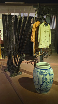 Antique display with a blue and white vase, Asian robe on a stand, spears, and a yellow jacket against a dark wooden background.
