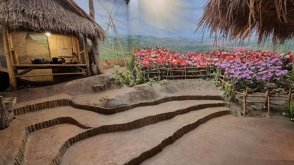 A rustic hut with a thatched roof near terraced soil beds. Vibrant red and purple flowers are fenced by bamboo, set against a scenic backdrop.