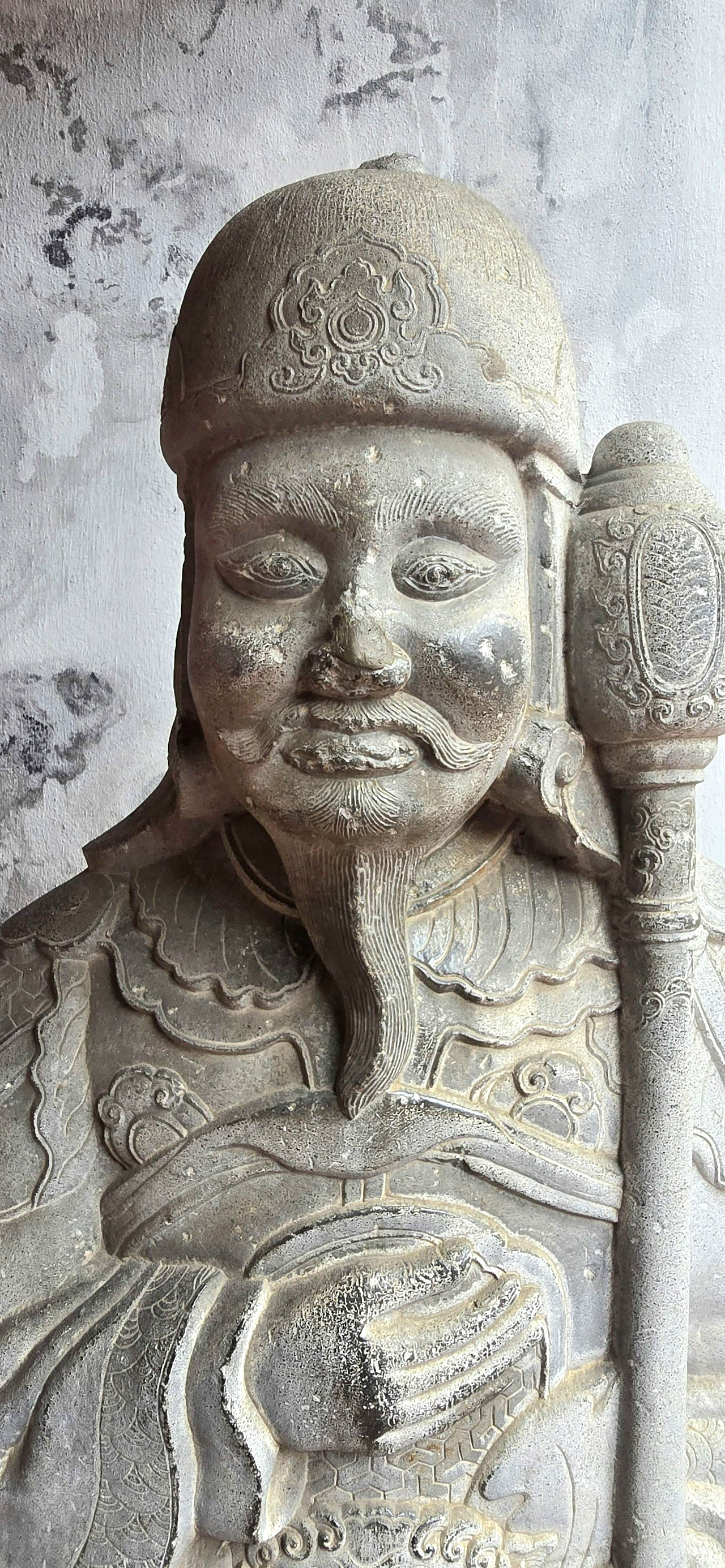 Stone statue of a figure with a helmet and intricate armor, holding a staff. The background features a textured, weathered wall.