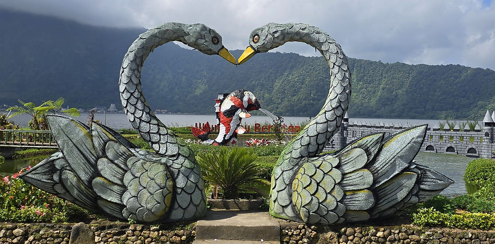 Two large swan sculptures form a heart in a lakeside garden, with mountains, cloudy sky, and a colorful fish statue behind.