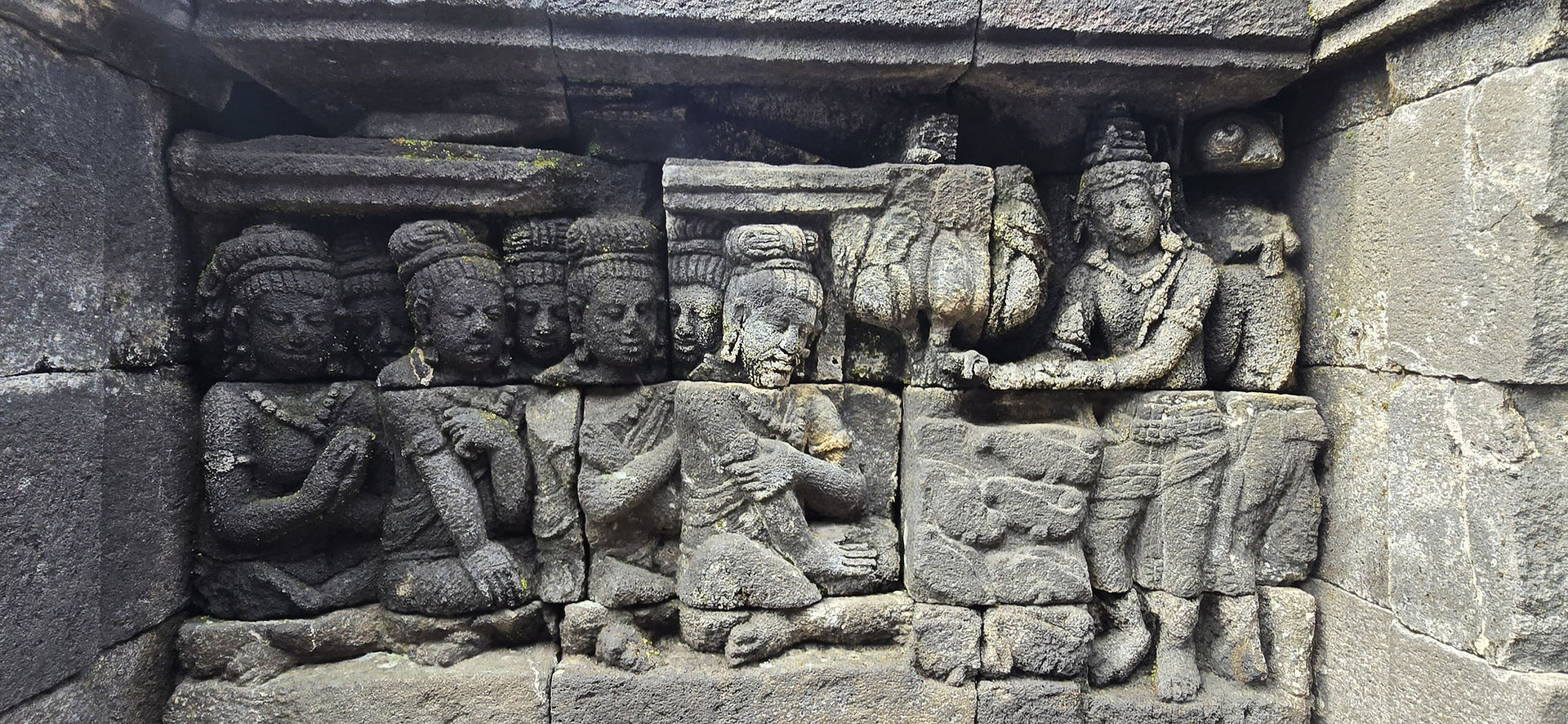 Stone relief of figures in intricate detail, featuring multiple people with headdresses in a scene, set on an ancient textured wall.