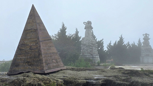 Foggy scene with a stone pyramid inscribed with text and two intricate statues in the background. Dense greenery surrounds the area.