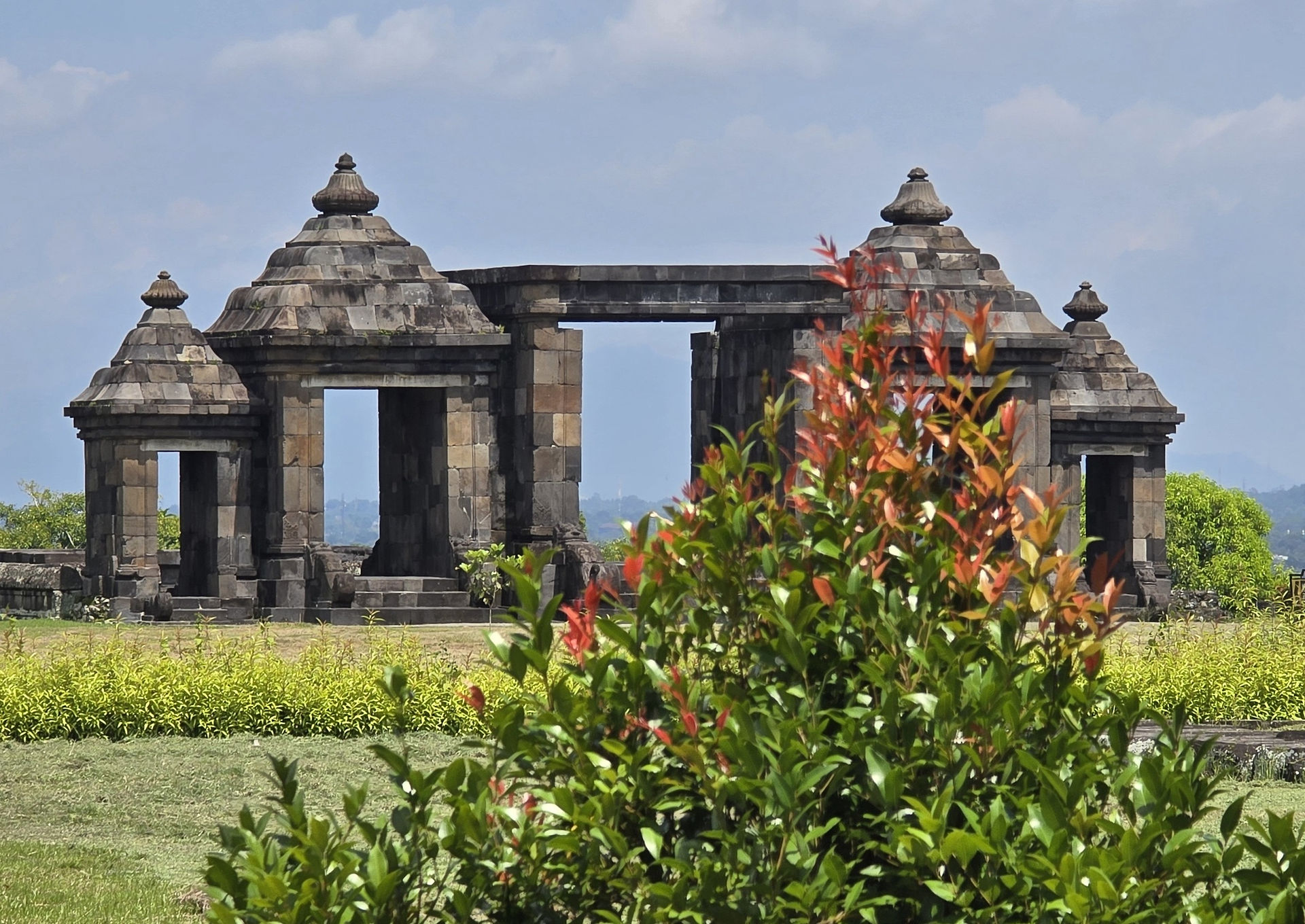 Ancient stone temple with three towers in the background, surrounded by lush greenery and vibrant red-tipped foliage under a cloudy sky.