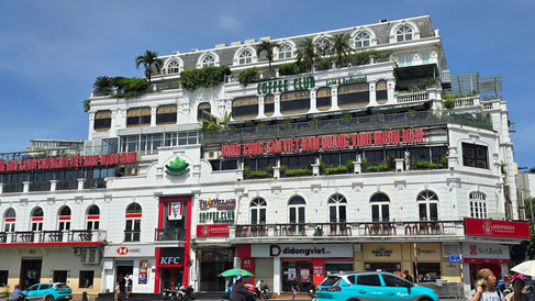 White building with green signage, red banners in Vietnamese, KFC and bank logos at street level, people, cars, and scooters nearby.