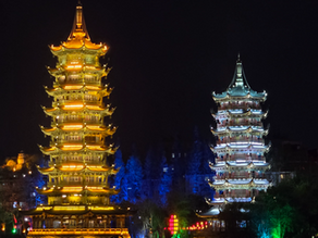 Two illuminated pagodas at night, one glowing gold, the other silver-blue, set against a dark sky. Colorful lights dot the surrounding trees.