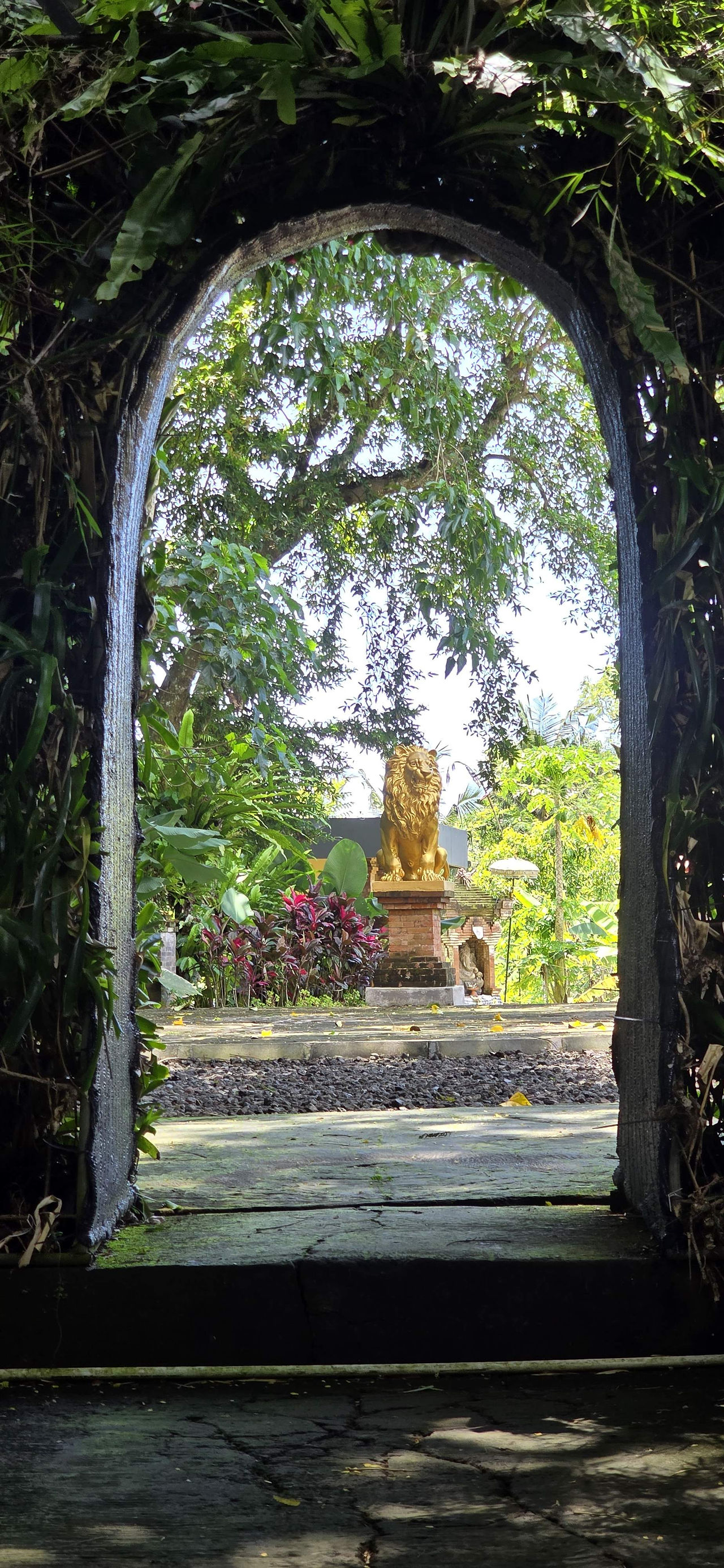 Golden lion statue on a stone pedestal, surrounded by lush greenery and red flowers, viewed through an archway covered in plants.