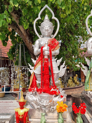Standing silver deity holding a sword, red sash over its shoulder, yellow and red flowers infront, trees in the background
