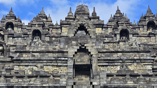 Stone temple facade with intricate carvings and Buddha statues set in niches. The weathered stone is gray and beige against a blue sky.
