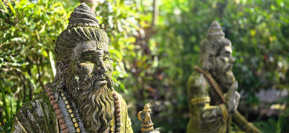 Two moss-covered stone statues of bearded figures in a lush garden setting. Both wear bead necklaces, with a serene and ancient appearance.
