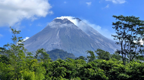 Mount Merapi volcano with a smoky peak looms behind lush green trees, set against a vivid blue sky with scattered clouds.