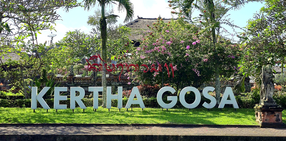 Kertha Gosa sign in a lush garden with colourful flowers, trees, and a stone statue. The setting is vibrant and peaceful.