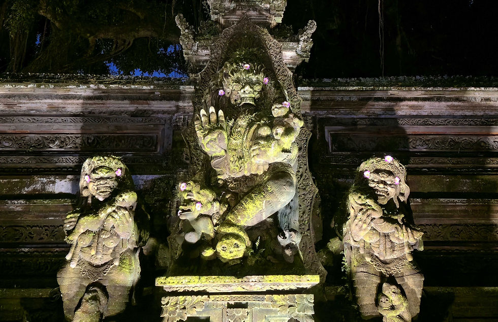 Three moss-covered stone statues with fierce expressions, adorned with pink flowers, stand against an ornate carved backdrop at night.