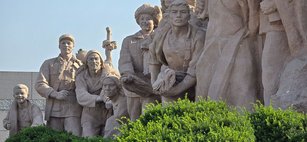 Stone sculptures of people in various poses stand in sunlight, surrounded by greenery. The background is a clear blue sky.