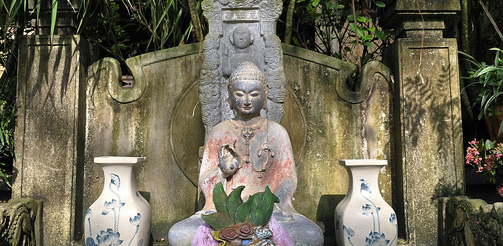 Stone Buddha statue in serene garden setting, flanked by ornate blue-and-white vases. Green foliage and weathered stone backdrop.
