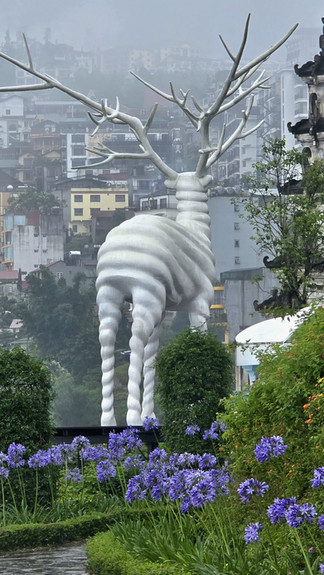 White twisted deer sculpture towers over a garden with purple flowers. Misty cityscape in the background adds a serene, foggy atmosphere.
