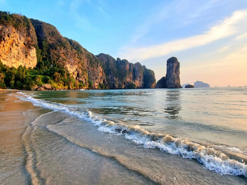 The coastline of Railay at sunset, with golden hues reflecting on the ocean and the silhouette of limestone cliffs. Small crashing waves on the golden beach