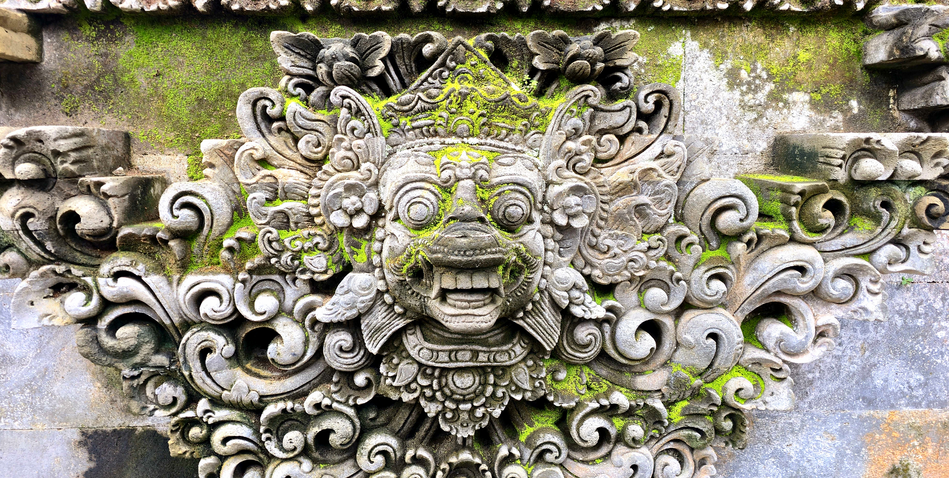 Ornate stone carving of a mythological face with intricate patterns and moss accents on a weathered surface, evoking an ancient, mystical aura.