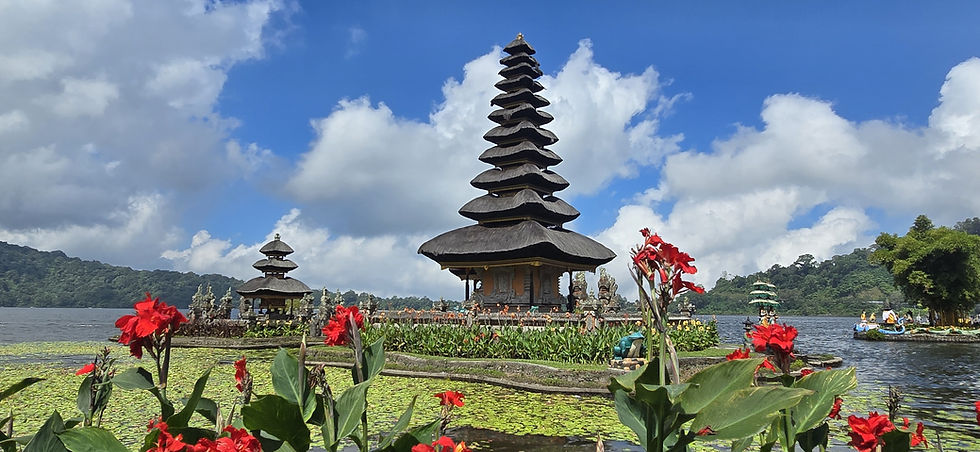 Balinese water temple with tiered pagodas, red flowers, lily-covered lake, and cloudy blue sky over green hills