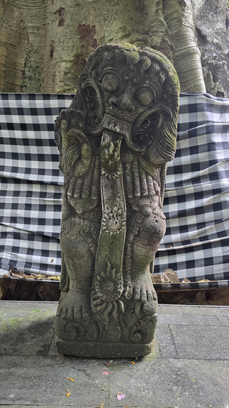 Moss-covered stone guardian statue with bulging eyes and tongue out, set before a black-and-white checkered cloth and tree