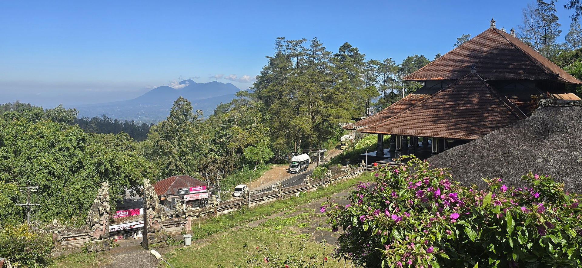Sunny mountain view with a Balinese temple roof, purple flowers, trucks on a hillside road, and clear blue sky.