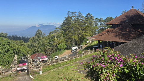 Sunny mountain view with a Balinese temple roof, purple flowers, trucks on a hillside road, and clear blue sky.
