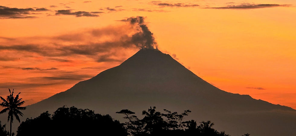 Volcano at sunset, emitting smoke against an orange sky. Silhouette of trees in the foreground creates a dramatic, serene scene.
