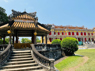 Ornate pavilion with curved steps in foreground, vibrant historical building with red shutters behind. Bright, sunny day, green shrubbery.