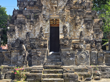 Ornate stone temple facade with carved figures and arched gates, centered dark doorway, sunlit greenery and potted flowers around steps