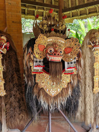 Three ornate traditional masks with elaborate designs and long hair, displayed indoors with a brick wall and lush green plants visible.