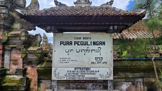 A sign at Pura Pegulingan temple with entry rules. Weathered stone architecture surrounds, set against a cloudy sky and green foliage.