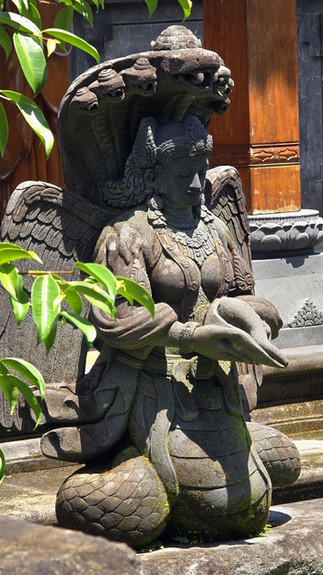 Stone statue of a mythical figure with wings and snake headdress in a garden setting, surrounded by green leaves and a carved stone background.