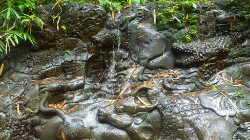 Stone carving of a reclining figure with intricate patterns, surrounded by animals, set in lush greenery with fallen leaves.