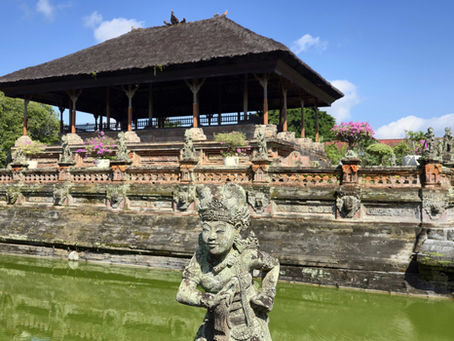 Stone statue before a traditional Balinese pavilion with lush greenery and vibrant flowers, reflected in a serene green pond under a blue sky.