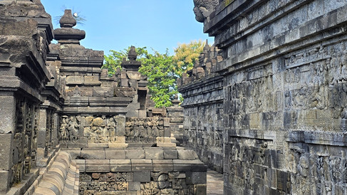 Ancient stone temple walls with intricate carvings against a blue sky. Greenery in the background, creating a serene, historic atmosphere.