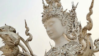 Ornate white statue with a serene face, intricate crown, and lotus flowers in hand. Detailed patterns and gold accents against a light sky.