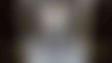 Ancient stone room with decorative wall carvings and 2 dark stone bed bases. Sunlight filters through a barred window. Mood is mysterious and dimly lit.