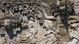 Weathered stone relief wall with carved faces and floral motifs, in gray and tan, on an ancient temple-like facade.