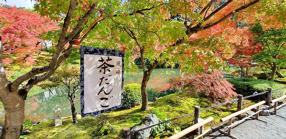 Japanese garden with colourful autumn leaves and a hanging sign with Japanese text. A serene pond and lush greenery create a peaceful scene.
